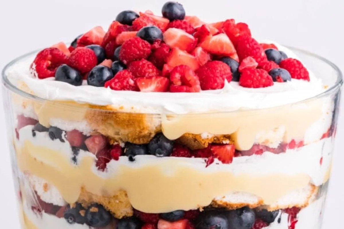 A patriotic dessert in a glass bowl featuring whipped cream, cake pieces, creamy filling, and assorted berries including strawberries, blueberries, and raspberries.