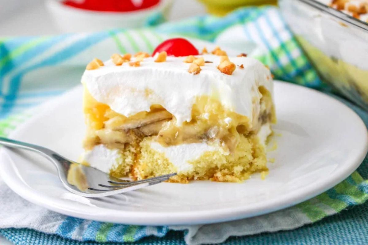 A slice of layered dessert with whipped topping, bananas, pudding, crushed nuts, and a cherry on top—one of those delightful shortcut desserts—served on a white plate with a fork on the side.
