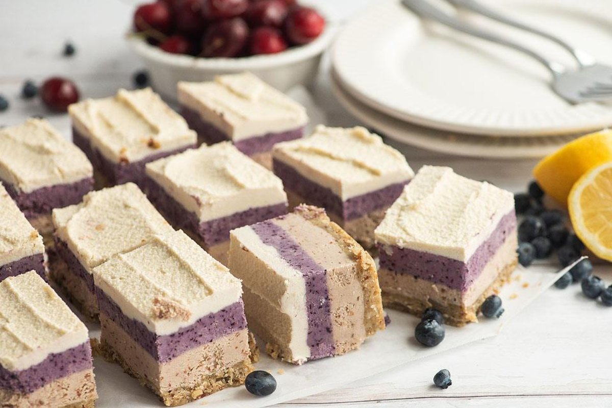 Slices of layered dessert bars with cream, purple filling, and a crust are arranged on a table. Blueberries, lemon wedges, a bowl of grapes, and a stack of plates with utensils complete the scene. Dive into cherry recipes for more delightful treats like these!