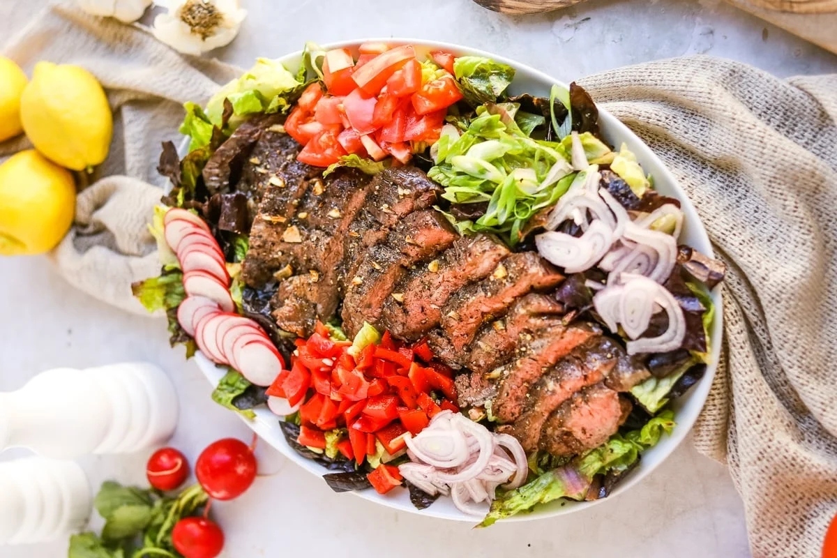 A salad made of mixed greens, sliced grilled flank steak, tomatoes, radishes, red peppers, shredded cheese, and red onions is presented on a white oval plate. Lemons and vegetables surround the dish, making it one of the most vibrant flank steak recipes you'll enjoy.