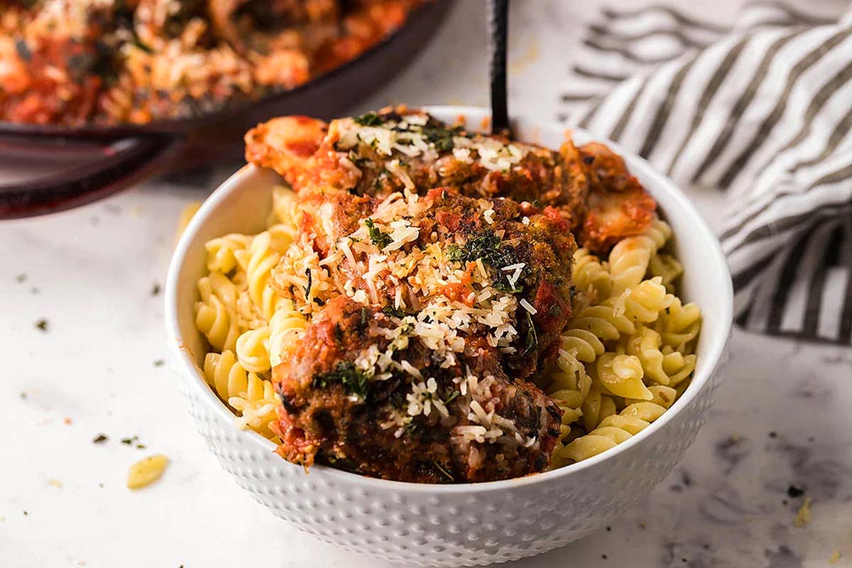 A bowl of fusilli pasta topped with savory meatballs and garnished with grated cheese and herbs, placed on a marbled surface next to a striped cloth, provides a delightful twist to traditional flank steak recipes.