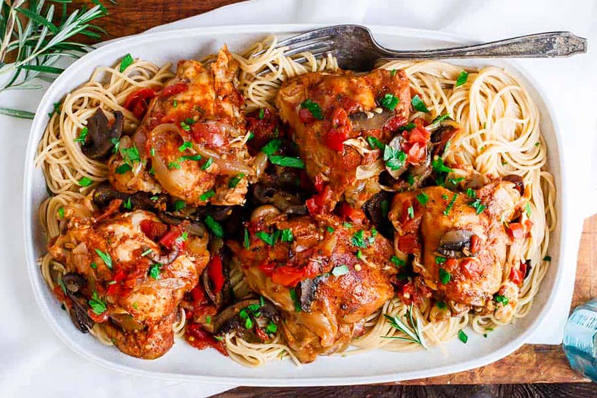 A dish of chicken cacciatore served over a bed of spaghetti, showcasing one of the best chicken thigh recipes, garnished with chopped herbs and diced red peppers on a white rectangular plate with a fork.