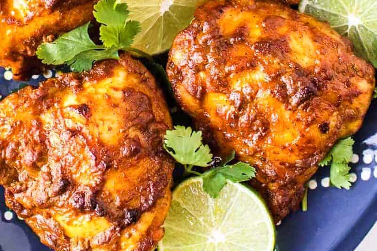 Close-up of three pieces of grilled chicken thighs with a golden brown crust, garnished with fresh cilantro and lime slices on a blue plate.