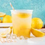 A plastic cup of iced fair lemonade with a straw, surrounded by fresh lemons, lemon slices, ice cubes, and paper straws, set against a blue background.
