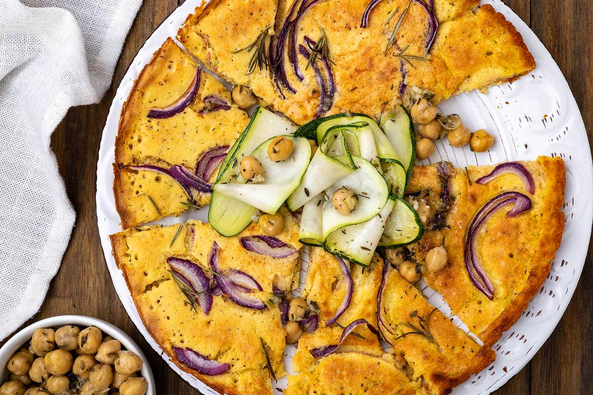 A round, sliced chickpea flatbread garnished with red onion slices, zucchini ribbons, rosemary, and chickpeas. A bowl of chickpeas is nearby, with a white cloth partially visible on the side. The flatbread is presented on a white plate on a wooden surface.