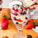 Four glasses of berry parfait filled with layers of coconut pudding, strawberries, and blackberries rest on a wooden board, surrounded by fresh strawberries and a halved coconut.