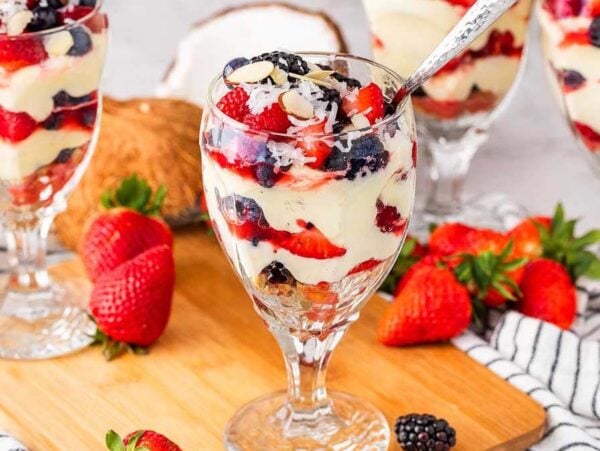 Four glasses of berry parfait filled with layers of coconut pudding, strawberries, and blackberries rest on a wooden board, surrounded by fresh strawberries and a halved coconut.