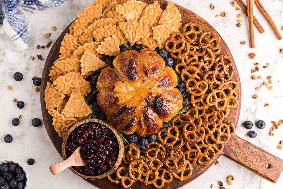 A round wooden board holds waffle cookies, pretzels, blueberries, and a pumpkin-shaped baked good with a bowl of blueberry sauce on the side&mdash;a perfect addition to your favorite blueberry recipes.