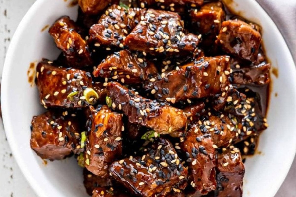 A white bowl filled with caramelized chunks of meat garnished with black and white sesame seeds and chopped green onions, showcasing one of the finest steak recipes.