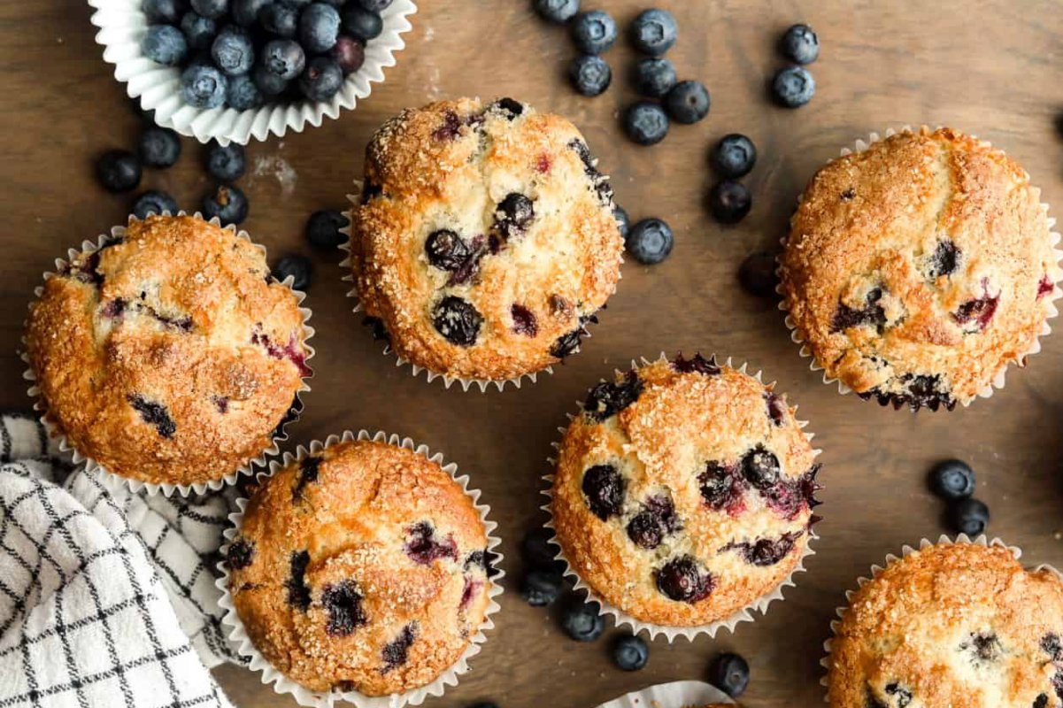 Six blueberry muffins are placed on a wooden surface with scattered blueberries and a checkered cloth nearby, showcasing one of the finest blueberry recipes.
