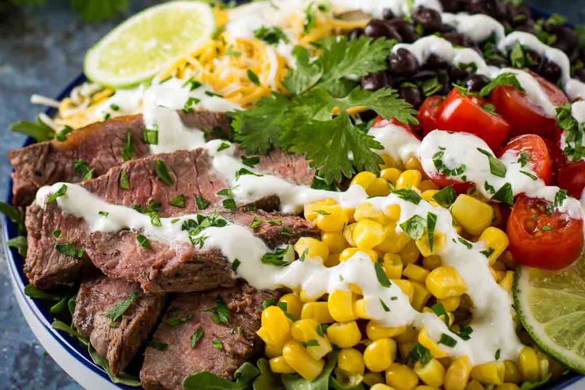 A colorful salad consisting of sliced steak, corn, cherry tomatoes, black beans, shredded cheese, and lime slices—all topped with a white creamy dressing and garnished with fresh cilantro. Perfect for those looking for vibrant steak recipes!