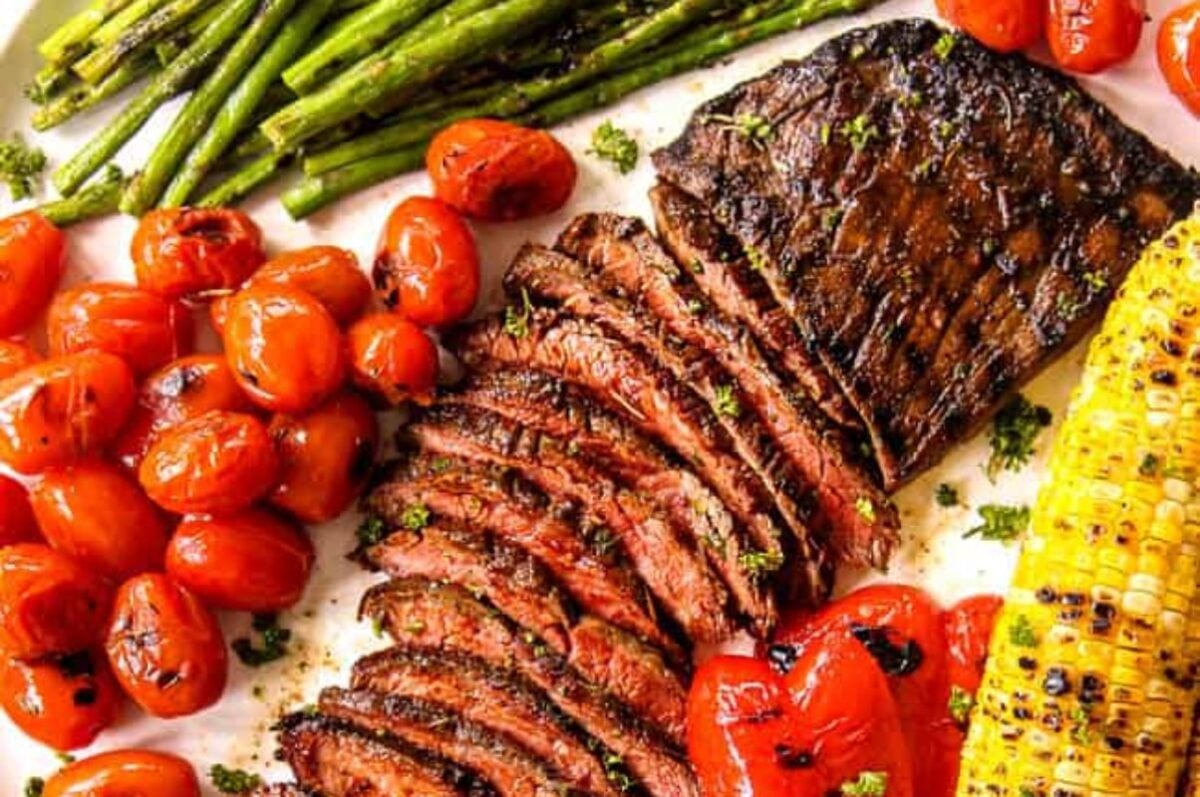 A platter of grilled flank steak recipes sliced, surrounded by roasted cherry tomatoes, asparagus, and grilled corn on the cob.