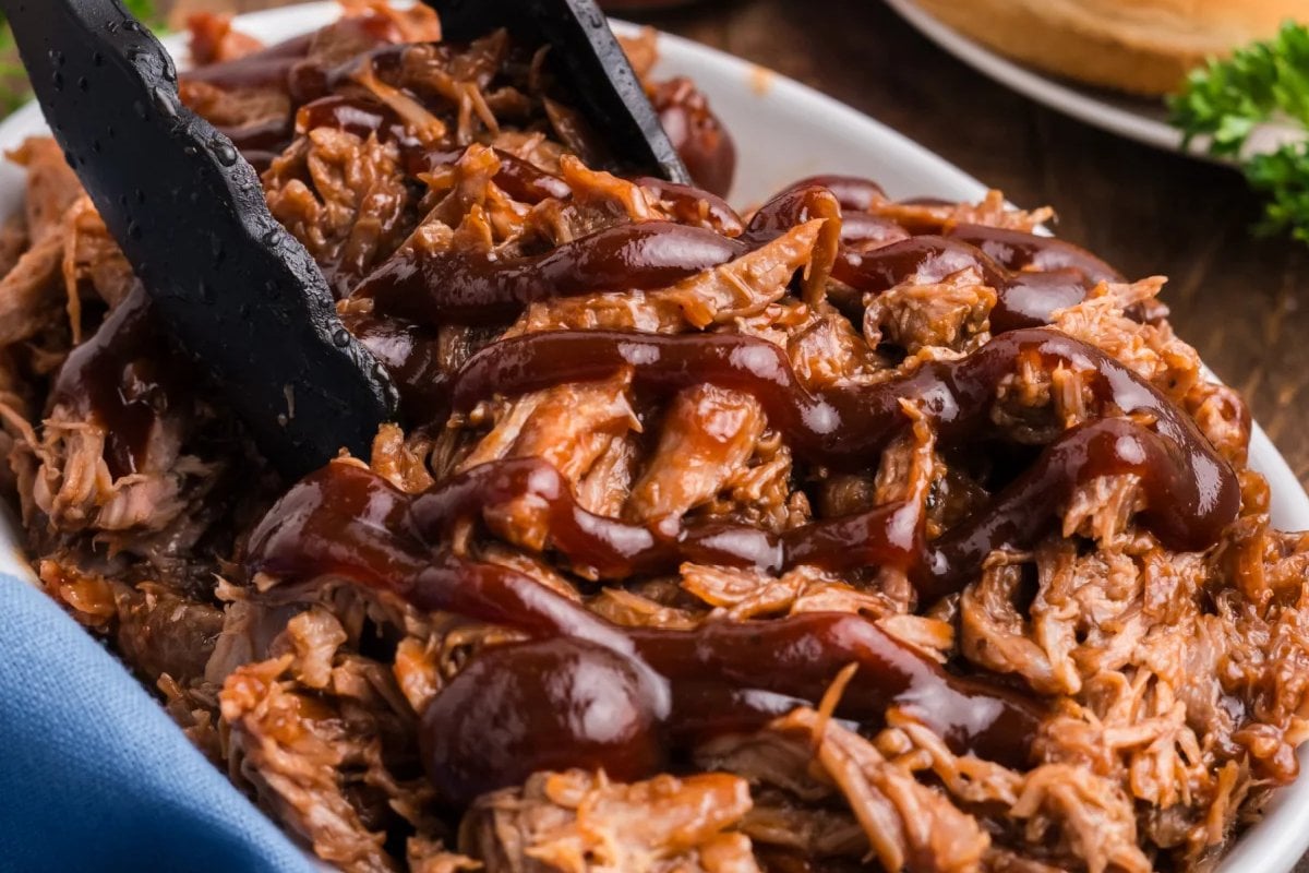 A close-up of a dish of pulled pork drizzled with barbecue sauce, showcasing one of the ultimate slow cooker dinners, with tongs resting on top.