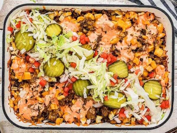 A casserole dish filled with a cheeseburger bake, topped with pickles, lettuce, tomato, and sauce—a delicious twist on classic big mac inspired recipes.