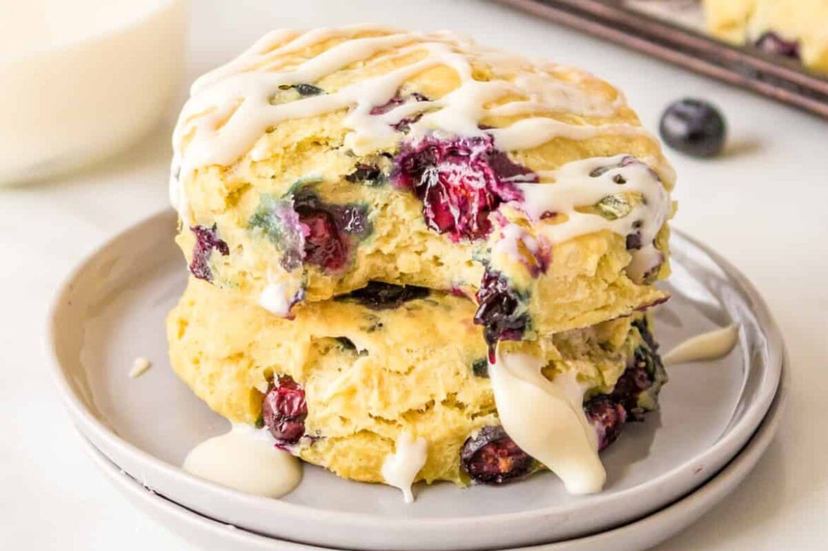 Two blueberry scones topped with white icing are stacked on a plate, showcasing one of the finest blueberry recipes. A baking sheet with more scones is visible in the background.