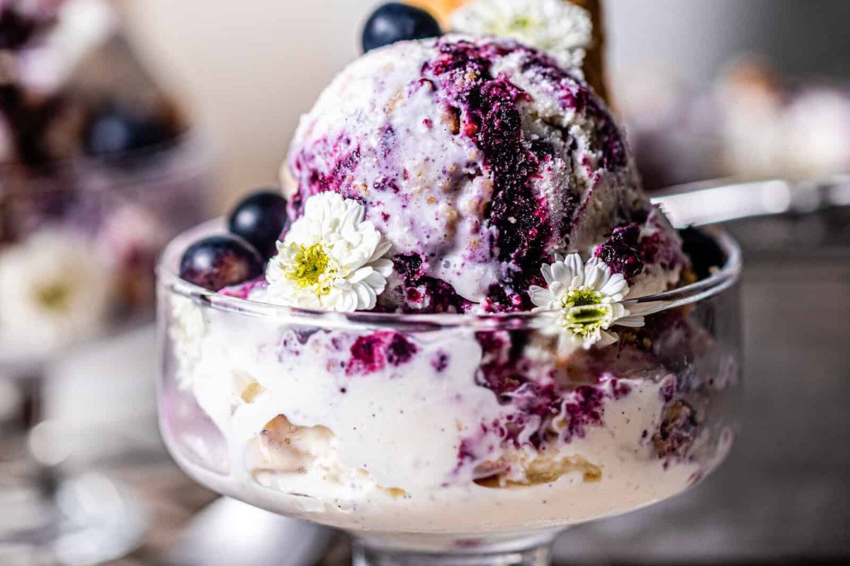 A close-up of a glass dessert dish holding scoops of blueberry swirl ice cream, garnished with small white flowers and blueberries, showcasing one of the finest blueberry recipes.
