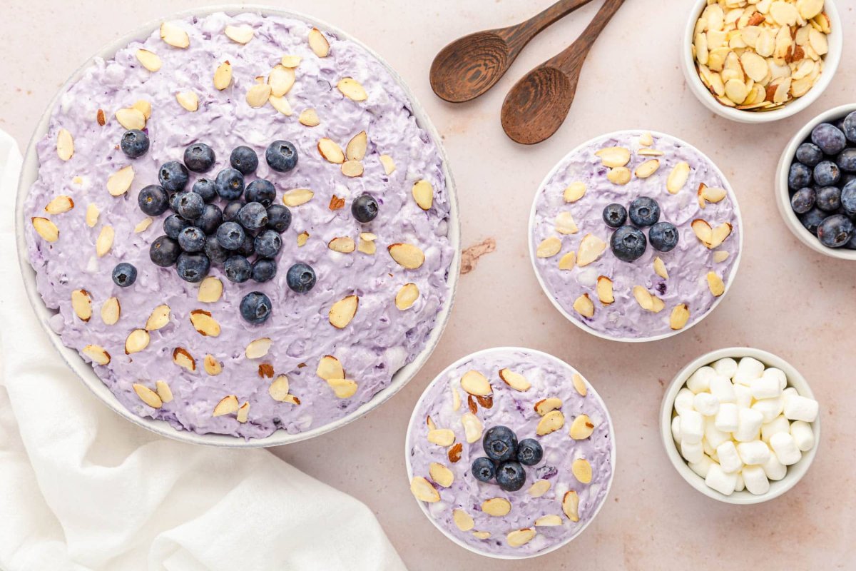 Bird's-eye view of a large bowl and two smaller bowls of purple dessert topped with blueberries and almond slices, showcasing a masterful touch in blueberry recipes. Additional bowls offer more blueberries, almond slices, and marshmallows, along with two wooden spoons.