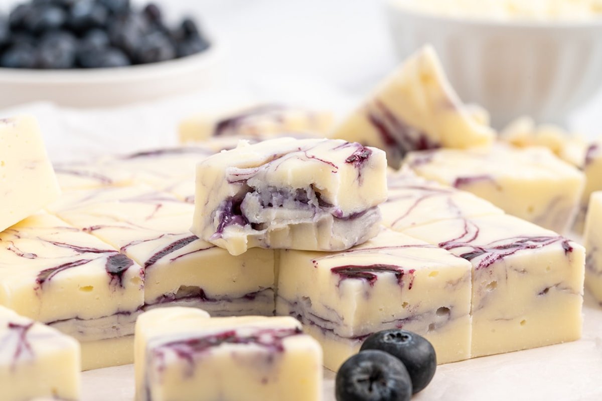 A close-up of white chocolate blueberry fudge pieces stacked on a surface, showcasing one of the finest blueberry recipes, with fresh blueberries and a bowl of fudge out of focus in the background.