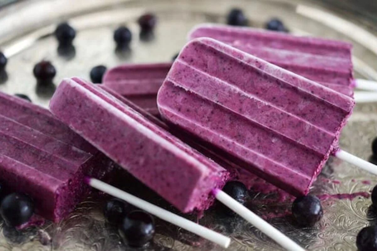 Four purple popsicles with visible berry specks are arranged on a silver tray, surrounded by scattered whole berries&mdash;an enticing addition to your blueberry recipes.