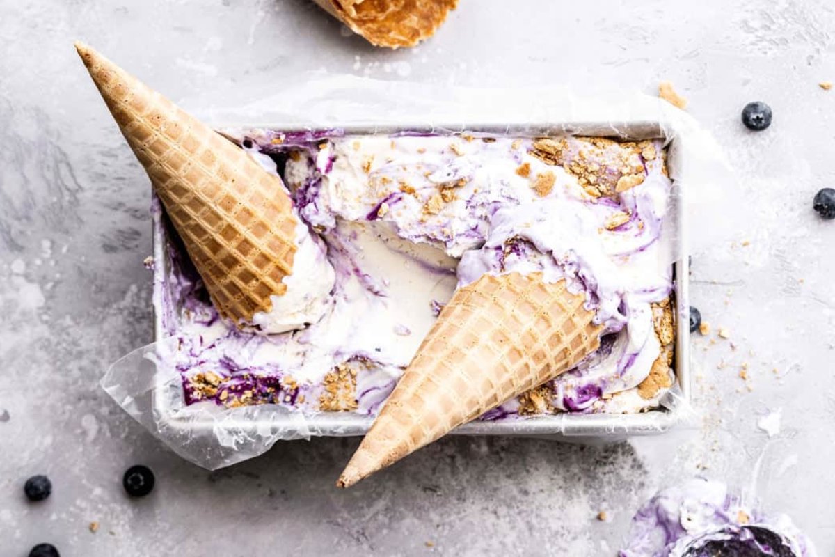A container of ice cream with a swirl pattern, topped with two waffle cones placed upside down, scattered pieces of waffle cone and blueberries around it&mdash;an irresistible addition to your favorite blueberry recipes.