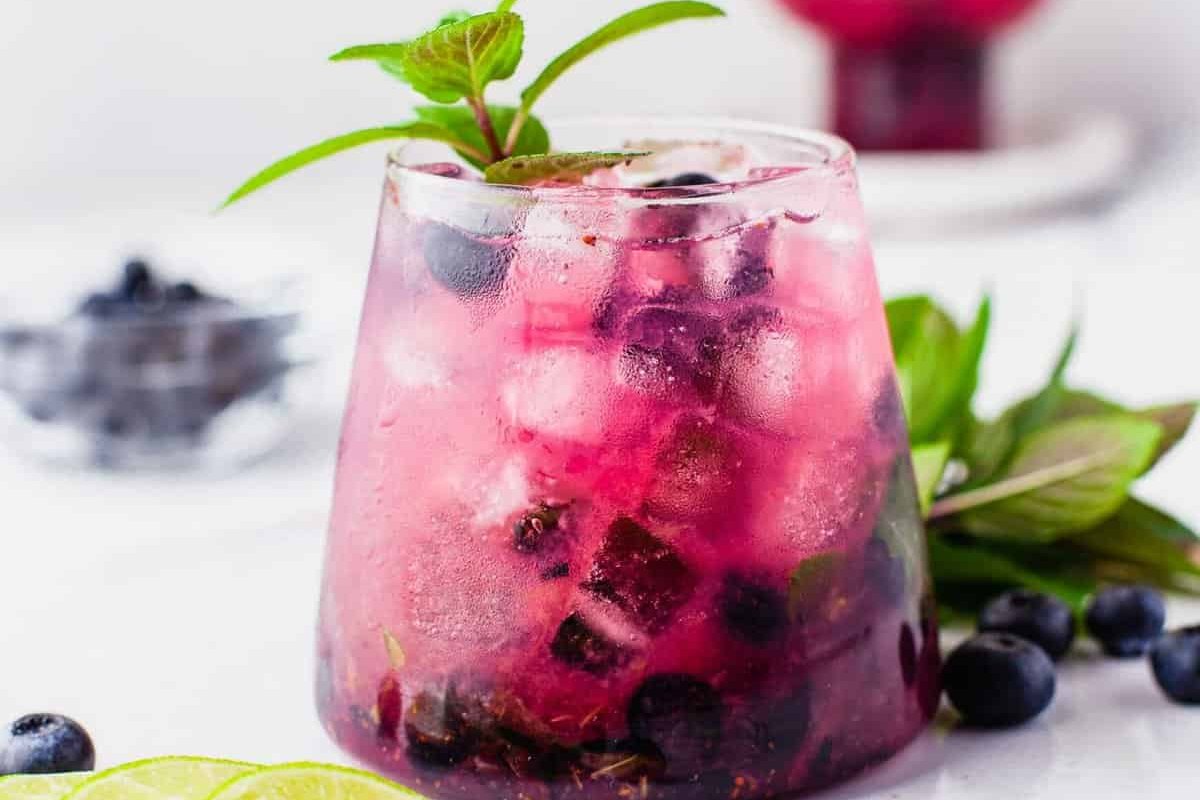 A glass of iced blueberry and mint drink, garnished with mint leaves, sits invitingly on a white surface. In the background, blueberries and lime slices hint at the refreshing ingredients used in this delightful addition to your favorite blueberry recipes.