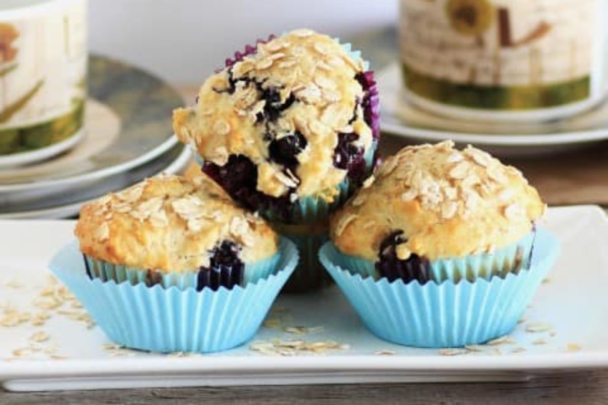 Three blueberry muffins in blue paper liners topped with oats on a white plate, showcasing one of the finest blueberry recipes, with a blurred background of two mugs and saucers.