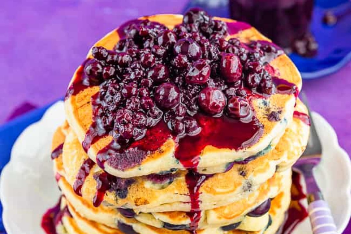 A stack of pancakes topped with a generous amount of blueberry compote sits on a white plate with a fork on the side, offering one of the finest delights in blueberry recipes.