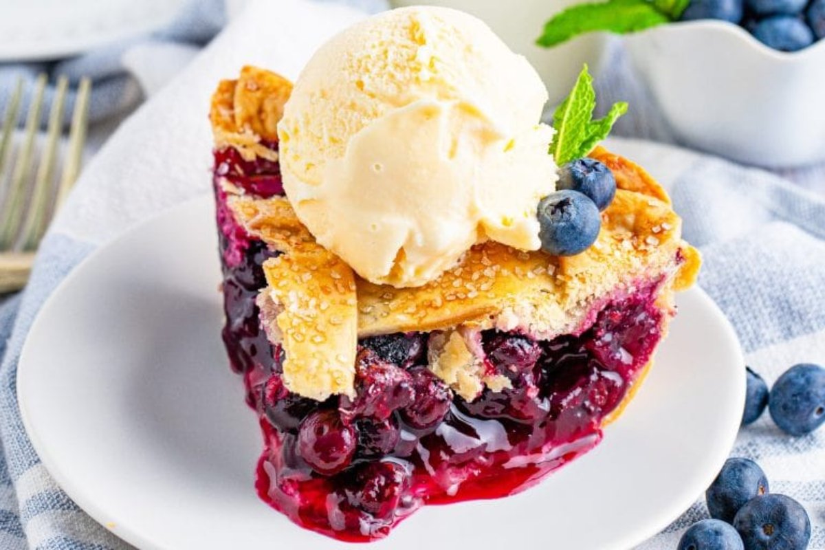 A slice of berry pie topped with a scoop of vanilla ice cream, garnished with fresh blueberries and a mint leaf, served on a white plate&mdash;an exquisite addition to your blueberry recipes collection.