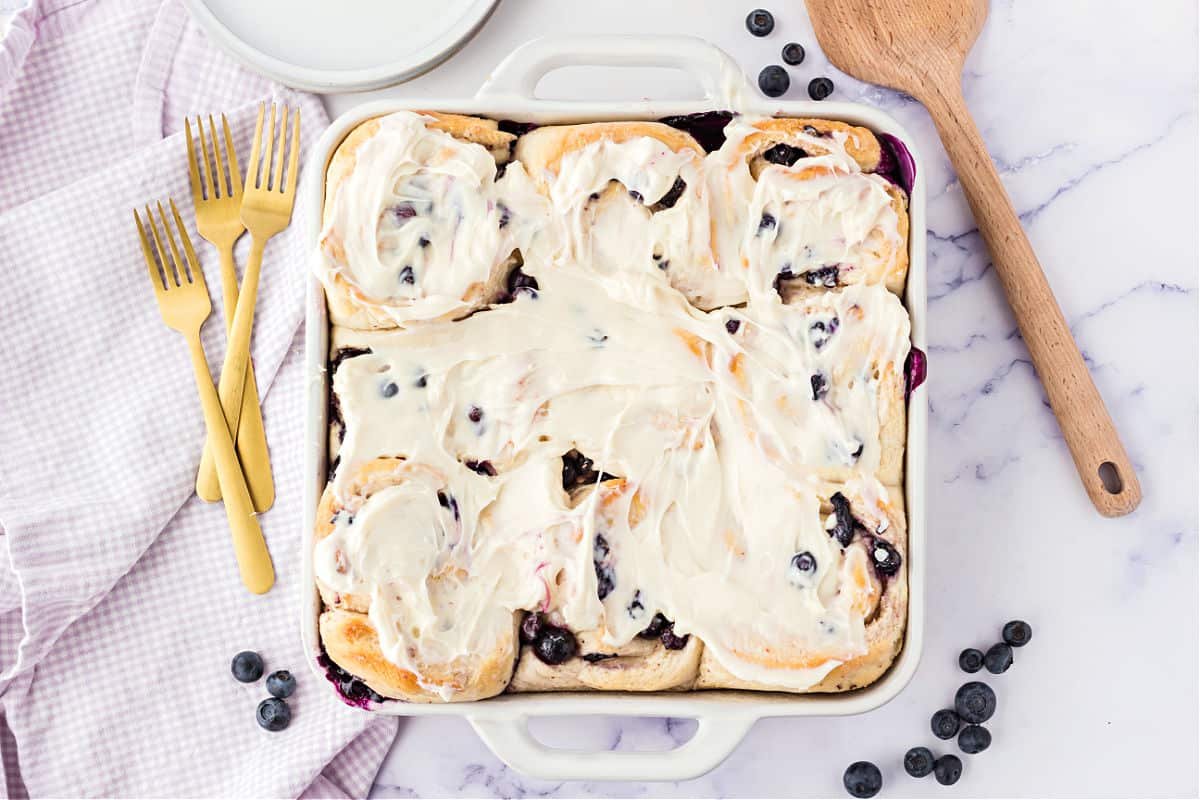 A baking dish filled with frosted blueberry cinnamon rolls, placed on a marble surface with scattered blueberries, a wooden spoon, a white cloth, and three gold forks nearby offers an enticing look at creative blueberry recipes.