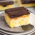 A slice of layered Boston cream poke cake with chocolate frosting on top is served on a small grey plate. A checked napkin, a spoon, and a bowl of custard are nearby.