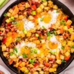 A hearty breakfast skillet containing a hash made of diced potatoes, bell peppers, onions, and melted cheese, topped with cooked eggs. Green onions are sprinkled on top as garnish.