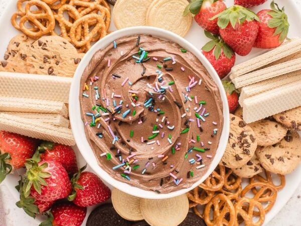 A platter of assorted snacks, including brownie batter dip with sprinkles, pretzels, strawberries, chocolate chip cookies, vanilla wafers, and wafer sticks, arranged on a white plate.