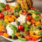A colorful burrata with tomatoes and pesto appetizer or salad featuring sliced tomatoes, mozzarella balls, olives, basil leaves, and pesto on a white plate with toasted bread and utensils in the background.