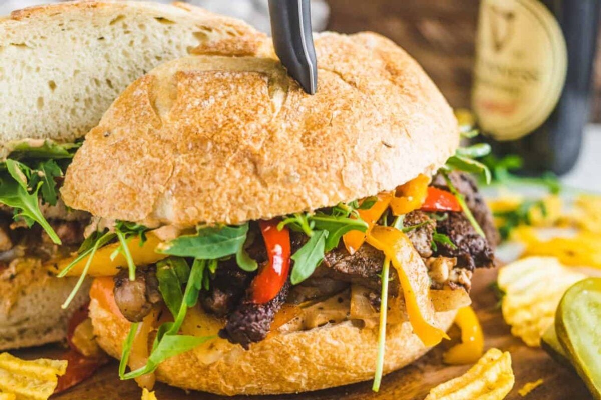 A crusty sandwich filled with grilled meat, arugula, sautéed peppers, and melted cheese, reminiscent of gourmet steak recipes, with a bottle and potato chips in the background.
