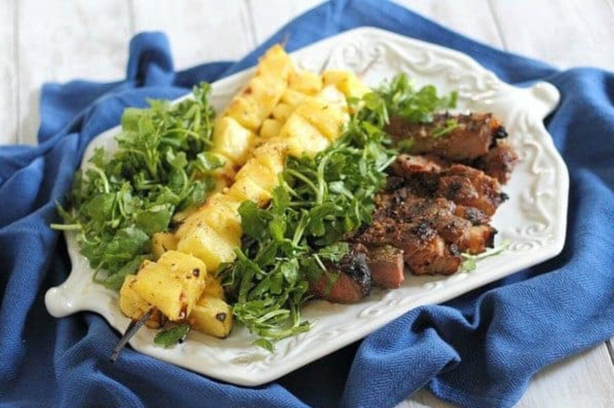 A white plate on a blue cloth showcases an appetizing ensemble: grilled meat, pineapple skewers, and a green leafy salad. Perfect for those seeking new steak recipes to elevate their culinary repertoire.