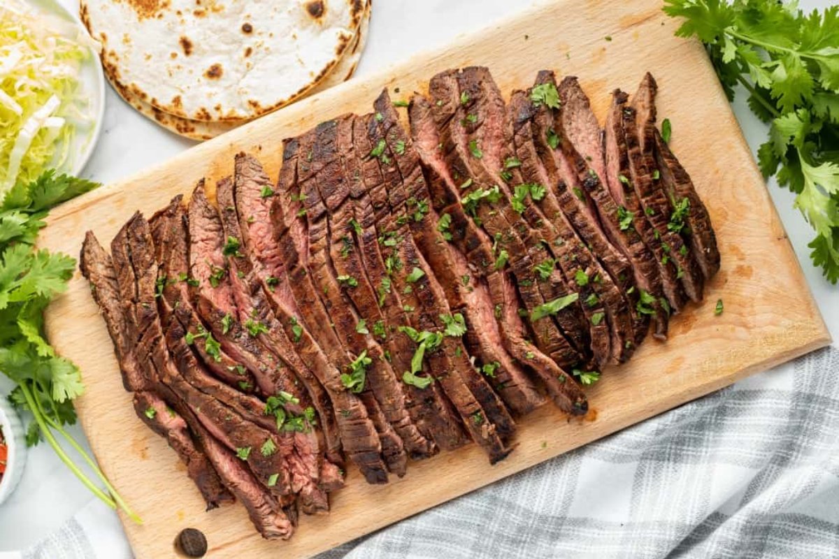Grilled beef slices garnished with chopped herbs on a wooden cutting board, this delightful dish is a highlight among steak recipes, accompanied by tortillas and greens in the background.