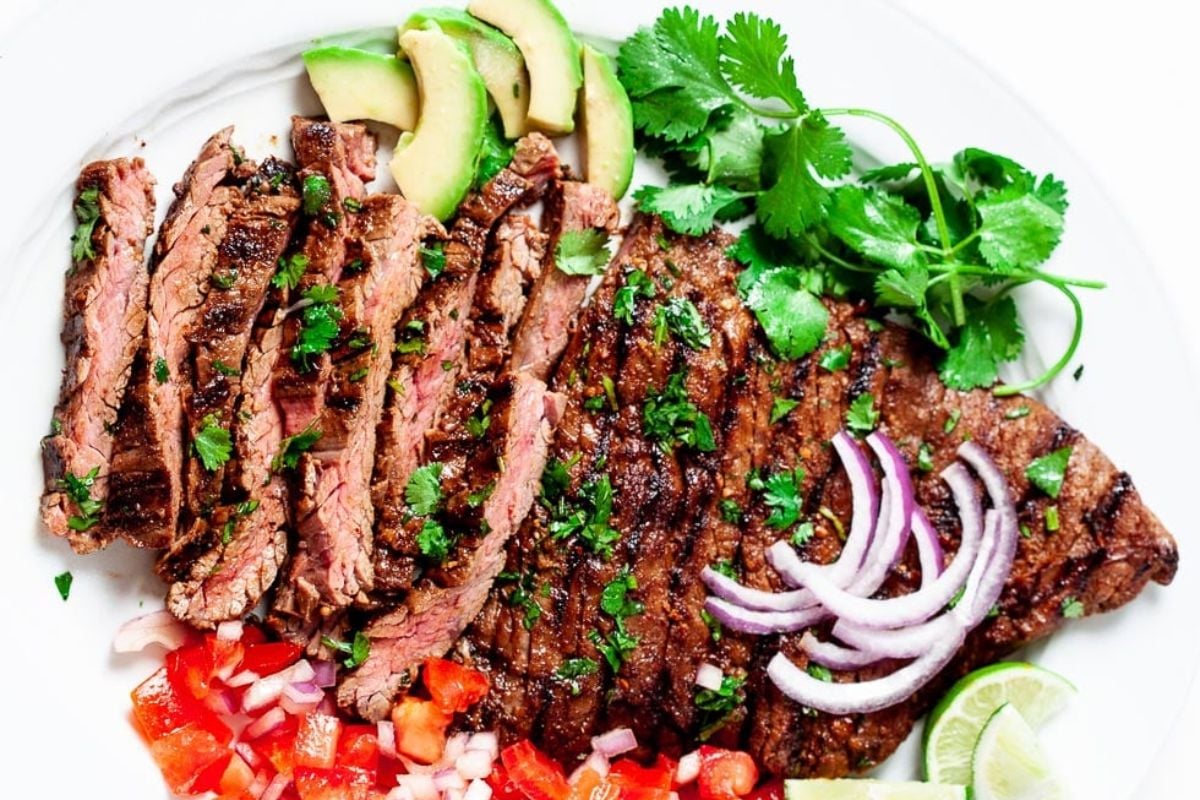 Grilled sliced steak garnished with cilantro, served with sliced avocado, red onion, diced tomatoes, and lime wedges on a white plate. Perfect for steak recipe enthusiasts.