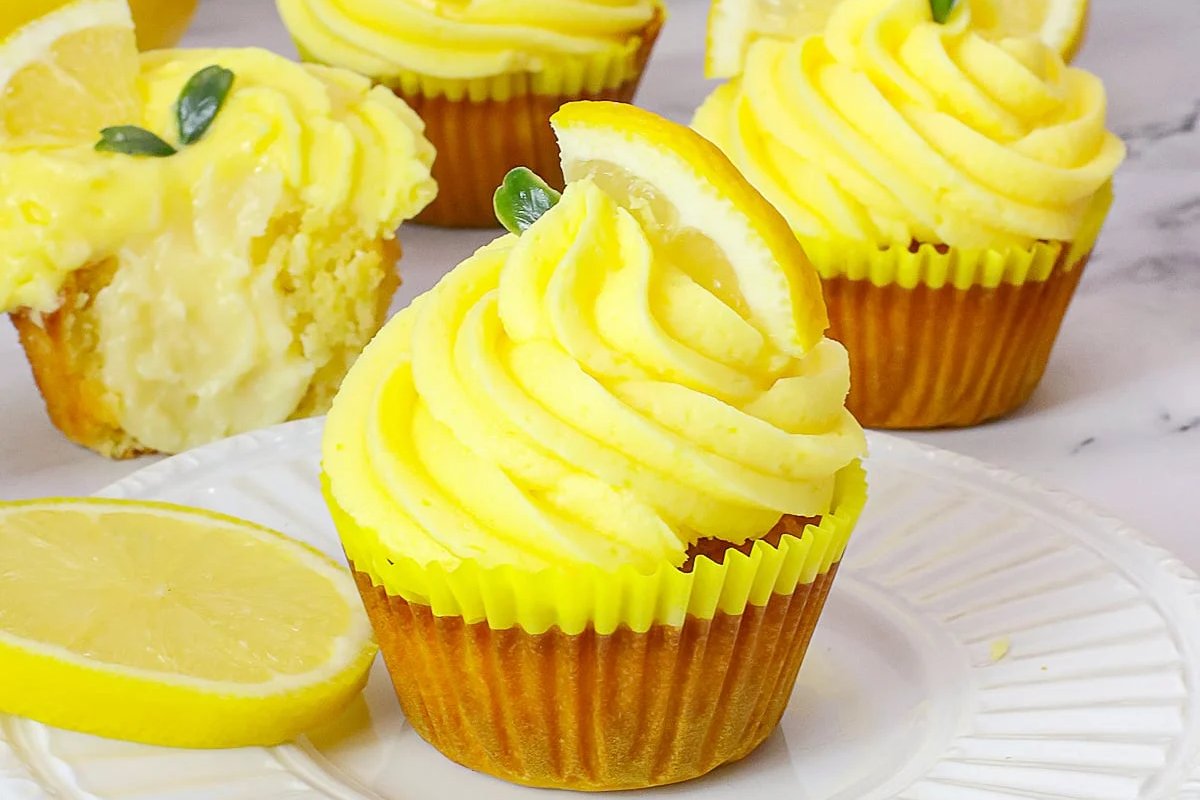 A plate with lemon-themed cupcakes topped with yellow frosting, lemon slices, and small green leaves. One cupcake is partially eaten, revealing a creamy filling. A lemon slice is also on the plate; truly irresistible lemon desserts.