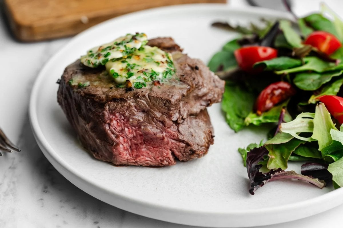 A cooked steak topped with herb butter on a white plate, served alongside a mixed green salad with cherry tomatoes, perfect for those seeking delicious steak recipes.