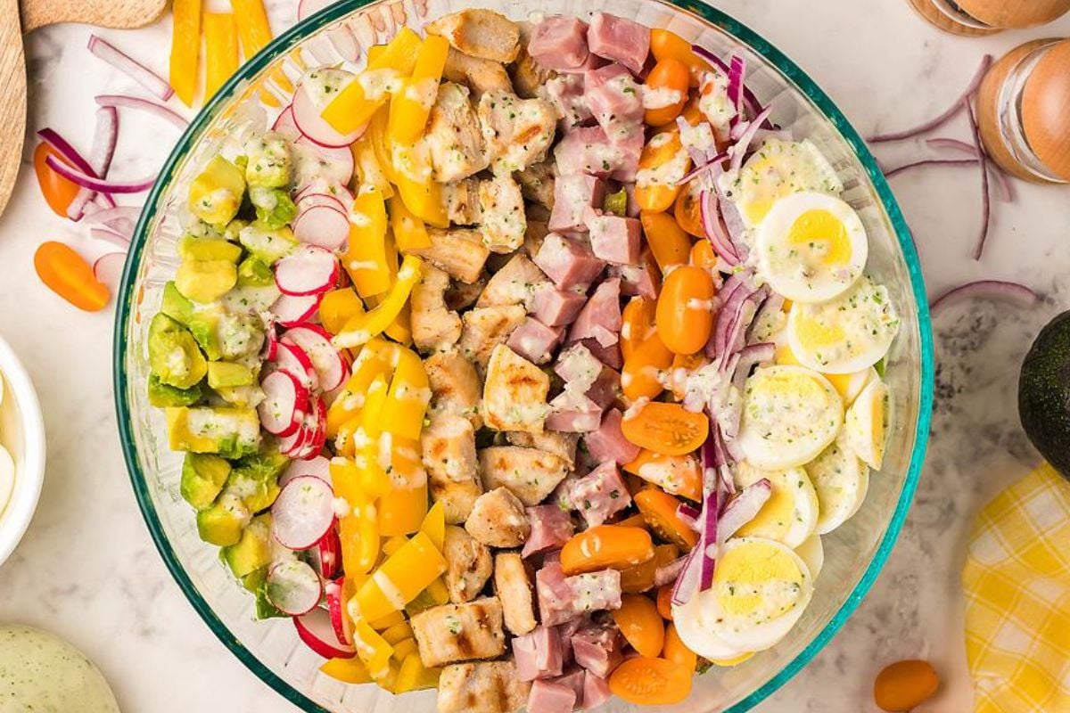 A clear bowl of chef salad with chopped ham, chicken, eggs, tomatoes, radishes, yellow peppers, pickles, red onions, and dressing.