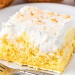 A slice of coconut cream poke cake topped with white frosting and sprinkled with toasted coconut served on a white plate with a fork beside it.