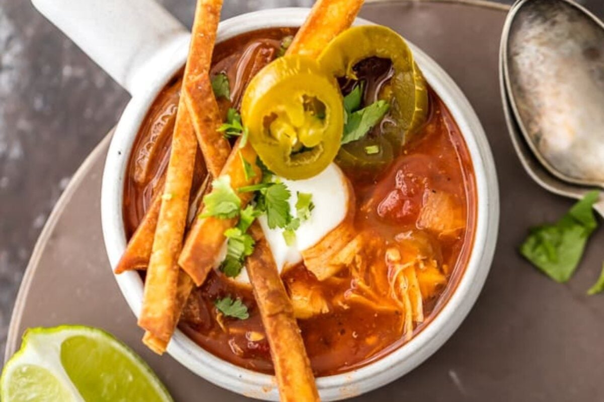 A white bowl filled with tortilla soup, slow-cooked to perfection, topped with sour cream, jalape&ntilde;os, and cilantro, served alongside a wedge of lime and a spoon.