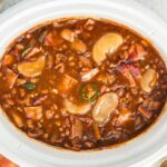 A white slow cooker filled with Slow Cooker Apple Pie Baked Beans. Nearby, there are sliced jalapeños, an apple slice reminiscent of slow cooker apple pie, and a wooden spoon.