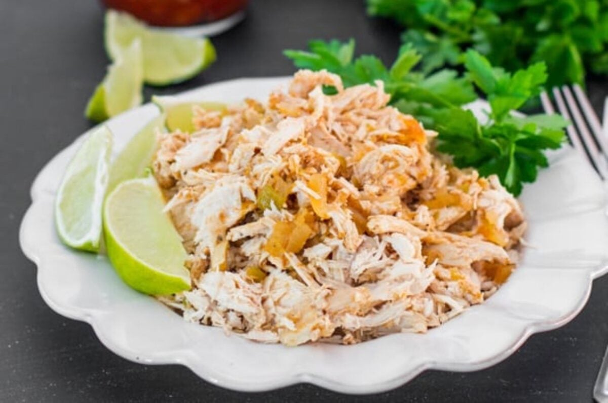 A plate of shredded chicken, perfectly cooked as one of those ideal slow cooker dinners, is served with lime wedges and garnished with parsley.