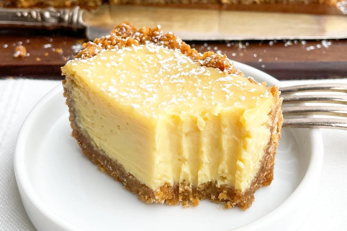 A slice of lemon cheesecake with a graham cracker crust on a white plate, lightly dusted with powdered sugar. A fork rests beside the plate.