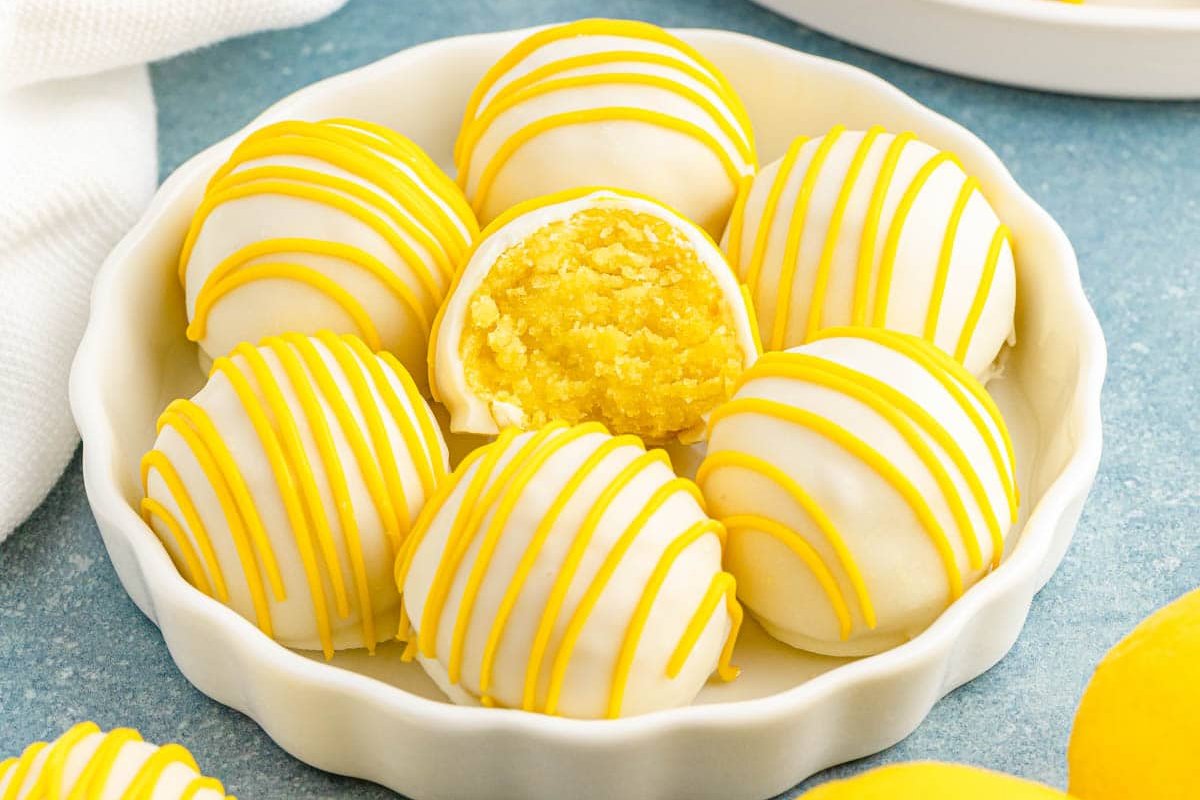 A plate of six round yellow and white cake truffles, with one truffle cut open to reveal a textured lemon filling. The truffles are drizzled with yellow icing, making these delightful lemon desserts a treat for the senses.