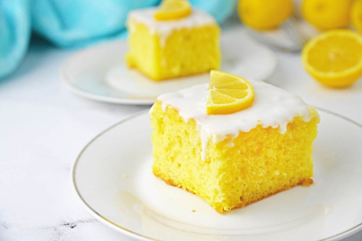 A slice of lemon cake with white icing and a lemon wedge on top sits on a white plate. Another slice and whole lemons are blurred in the background, showcasing this delightful addition to the world of lemon desserts.