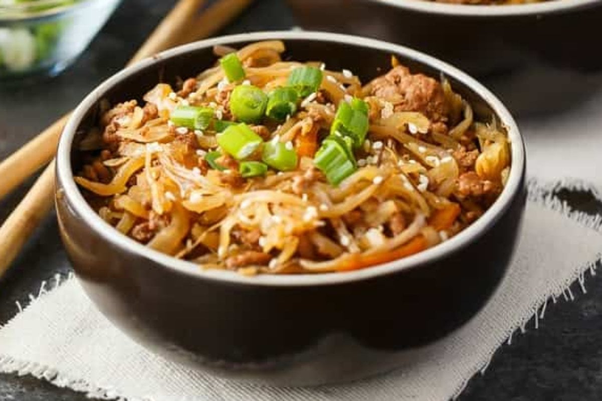 A bowl of stir-fried noodles topped with chopped green onions and sesame seeds, evocative of the rich flavors found in slow cooker dinners, placed on a napkin with chopsticks partially visible in the background.