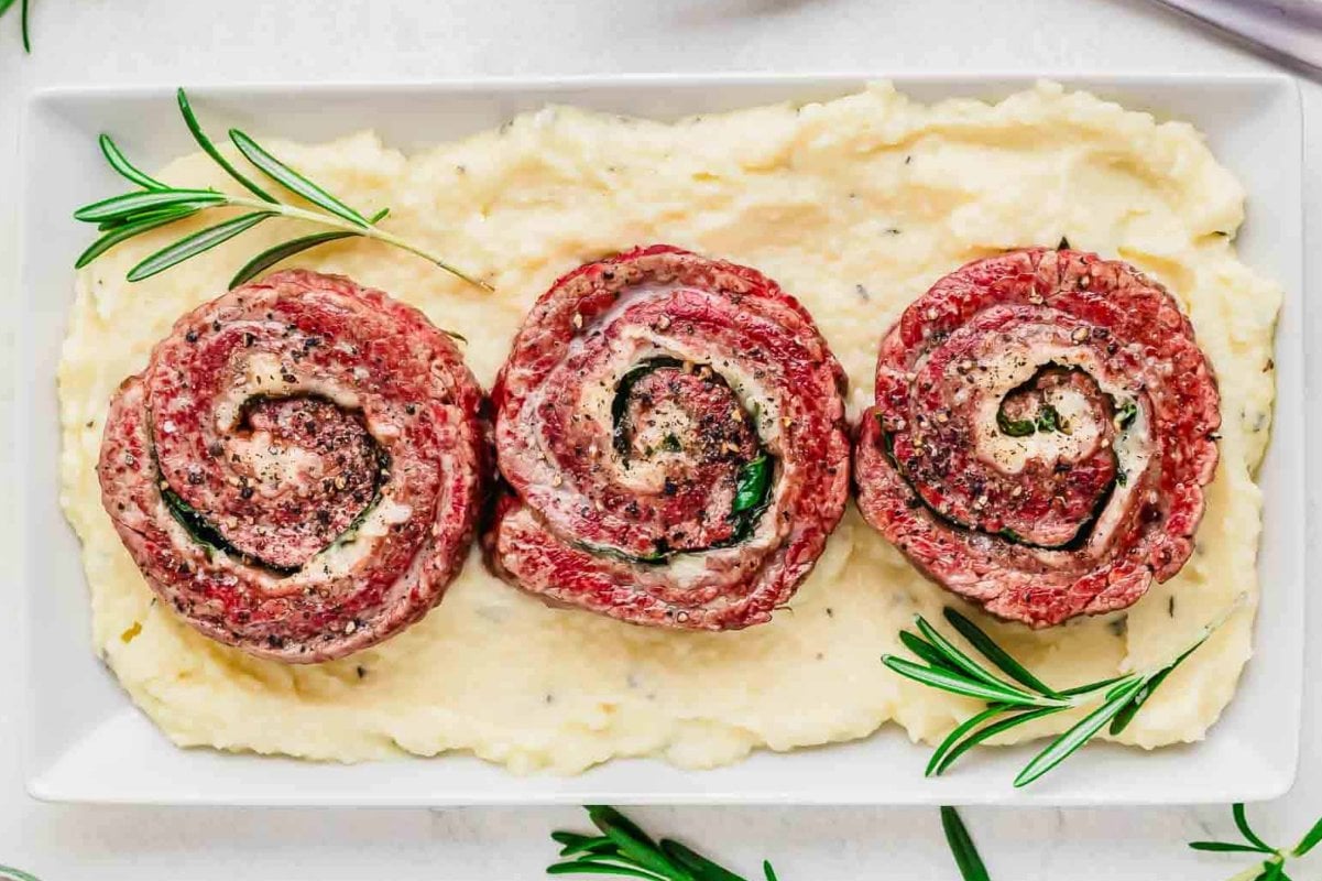 Three rolled beef steaks with herb stuffing sit on a bed of mashed potatoes, garnished with fresh rosemary sprigs on a white rectangular plate—an ideal addition to your collection of steak recipes.