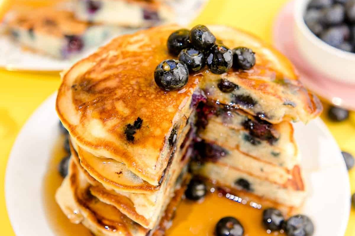 A stack of blueberry pancakes topped with fresh blueberries and drizzled with syrup on a white plate is a perfect example of delightful blueberry recipes.